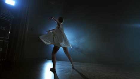 The-zoom-camera-moves-the-woman-dancer-across-the-stage-with-software-and-smoke.-A-modern-dramatic-ballet-a-woman-in-a-white-dress-spins-on-one-leg-and-jumps..